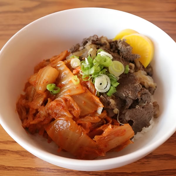 A white bowl of Japanese beef and kimchi over rice, garnished with green onions and yellow pickled daikon.