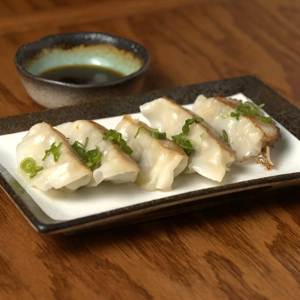 Pan-fried gyoza dumplings with green onions on a rectangular plate, with a small bowl of soy sauce.