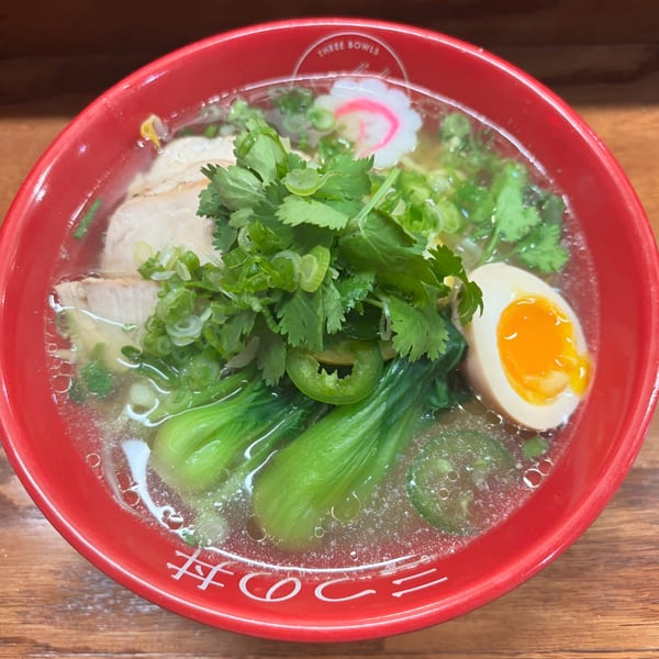 A vibrant red bowl of Three Bowls Ramen, featuring pork, bok choy, cilantro, a soft-boiled egg, and narutomaki.
