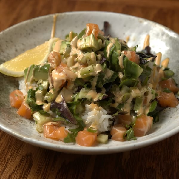 A Three Bowls Ramen poke bowl with salmon, avocado, greens, and sauce, served with a lemon wedge.