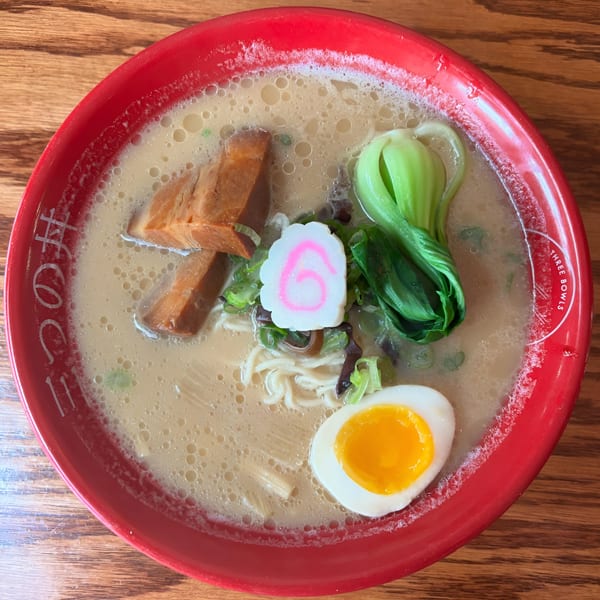 A red bowl of Three Bowls Ramen with pork, bok choy, fish cake, and a soft-boiled egg.