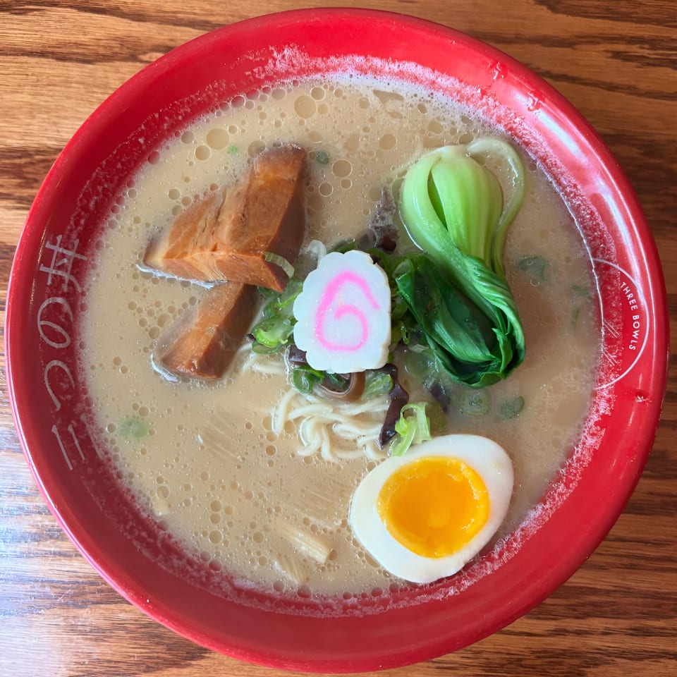 A red bowl of Three Bowls Ramen with pork, bok choy, fish cake, and a soft-boiled egg.
