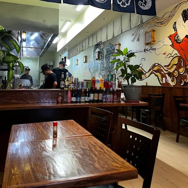Three Bowls Ramen interior with chefs behind the counter, Japanese wall art, and empty wooden tables.